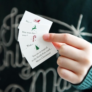 May include: A hand holding a stack of white Christmas cards with festive illustrations and text. The cards feature a sleigh, reindeer, candy cane, and Christmas tree. The text includes holiday wishes and the phrase "Merry Christmas!"