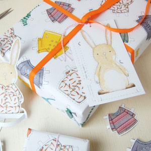 Puede incluir: Papel de regalo blanco con una ilustraci&oacute;n de un conejo y una etiqueta que dice "Para ti, de m&iacute;!" El papel presenta un patr&oacute;n repetitivo de mu&ntilde;ecas de papel que llevan diferentes atuendos, incluyendo un impermeable amarillo, un su&eacute;ter azul y una camisa con zanahorias. El regalo est&aacute; atado con una cinta naranja.