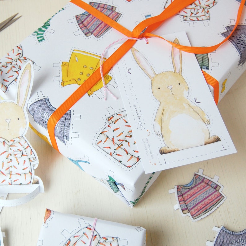 Puede incluir: Papel de regalo blanco con una ilustraci&oacute;n de un conejo y una etiqueta que dice "Para ti, de m&iacute;!" El papel presenta un patr&oacute;n repetitivo de mu&ntilde;ecas de papel que llevan diferentes atuendos, incluyendo un impermeable amarillo, un su&eacute;ter azul y una camisa con zanahorias. El regalo est&aacute; atado con una cinta naranja.
