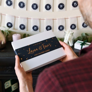 A white advent calendar box with a navy blue sleeve around it and foiled wording is being held. Rows of hung envelopes are on the wall behind it.