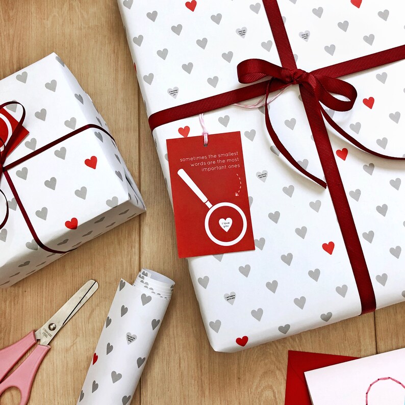 Tiny Heart Valentines Gift Wrap. White gift wrap featuring tiny grey and red hearts, some with tiny messages in is used to wrap a gift and finished off with a red bow.