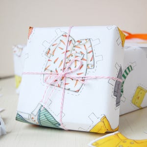 Puede incluir: Una caja de regalo blanca envuelta en papel con una ilustraci&oacute;n de dibujos animados de una camisa con un patr&oacute;n de zanahorias. La caja est&aacute; atada con un lazo de cuerda rosa y blanca.
