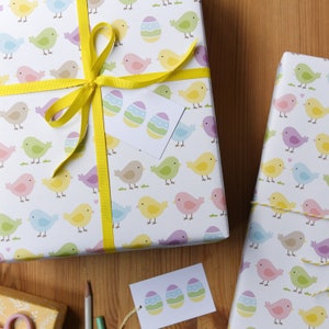 May include: Two gift boxes wrapped in white paper with a repeating pattern of colorful birds and Easter eggs. The boxes are tied with yellow ribbon and have gift tags with a similar pattern.