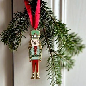 A gold, red, and green enamel Nutcracker Christmas tree decoration hangs from a red ribbon on a door handle, in front a Christmas tree branch.
