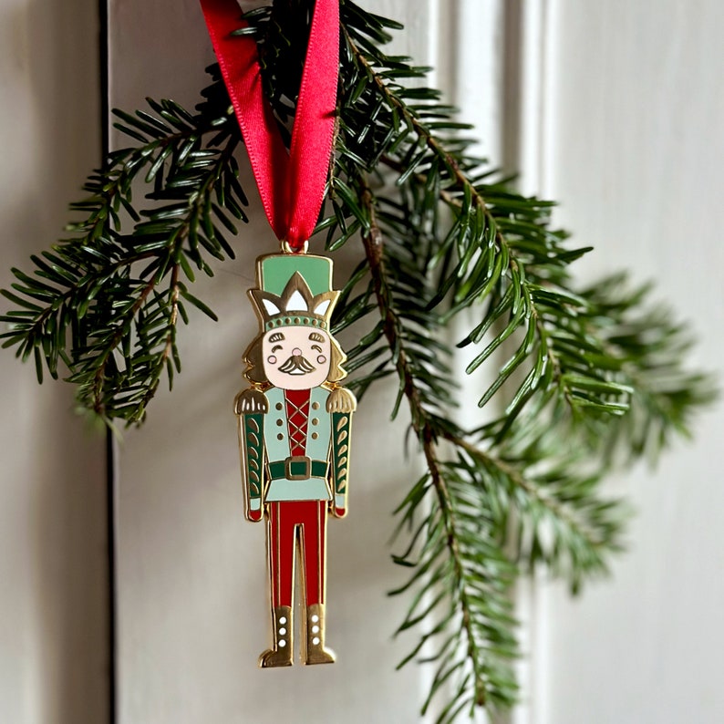 A gold, red, and green enamel Nutcracker Christmas tree decoration hangs from a red ribbon on a door handle, in front a Christmas tree branch.