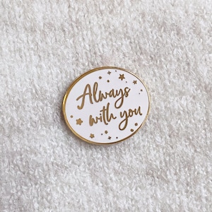 White and gold oval pin badge reading always with you in a joined up handwriting font. The gold rimmed white badge is attached to a cream coloured fine knit jumper