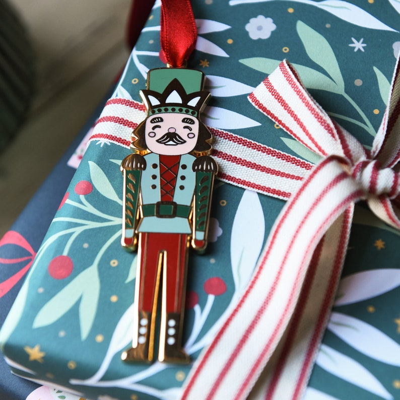 A gold, red, and green enamel Nutcracker Christmas tree decoration hangs from a red ribbon on a door handle, on a pile of wrapped Christmas gifts.