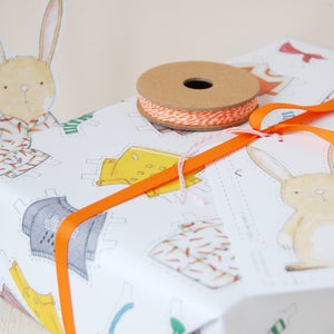 Puede incluir: Papel de regalo blanco con un patr&oacute;n repetido de recortes en forma de conejo que llevan diferentes atuendos. El papel est&aacute; envuelto alrededor de una caja de regalo y atado con una cinta naranja y un carrete de hilo naranja y blanco.