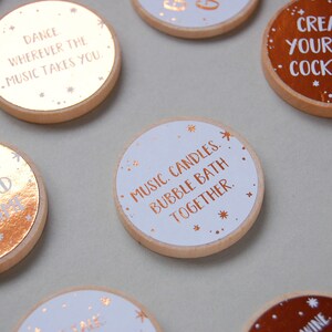 15 wooden disks scattered on a grey background. 7 of the wooden disks have white stickers with copper foiled writing on them, 8 of the disks have copper stickers with white writing on them