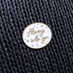 White and gold oval pin badge reading always with you in a joined up handwriting font. The gold rimmed white badge is attached to a grey coloured chunky knit jumper