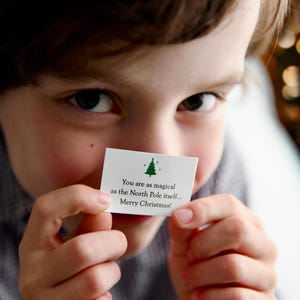 May include: A child holds a white card with a green Christmas tree graphic. The card reads, "You are as magical as the North Pole itself... Merry Christmas!" The child's eyes are visible, and the background is blurred.
