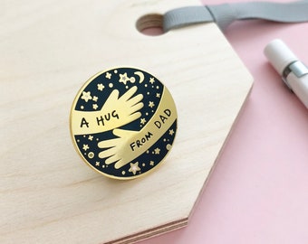 May include: A gold-colored enamel pin with a black background. The pin features two hands in a hug with stars and a crescent moon. The text "A HUG FROM DAD" is written around the hands.