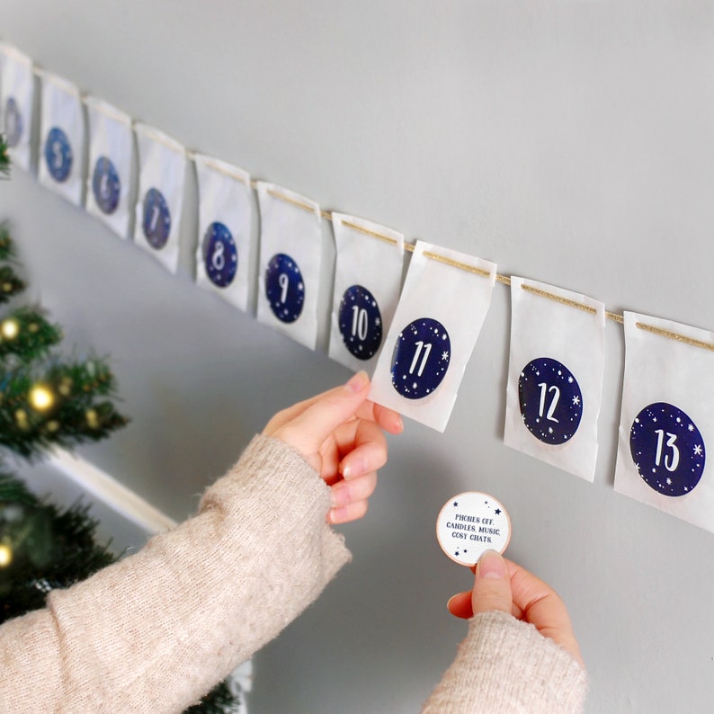 A row of white envelopes, each with a navy blue numbered sticker on, is hung on grey wall. One hand holds an envelopes and a wooden token.