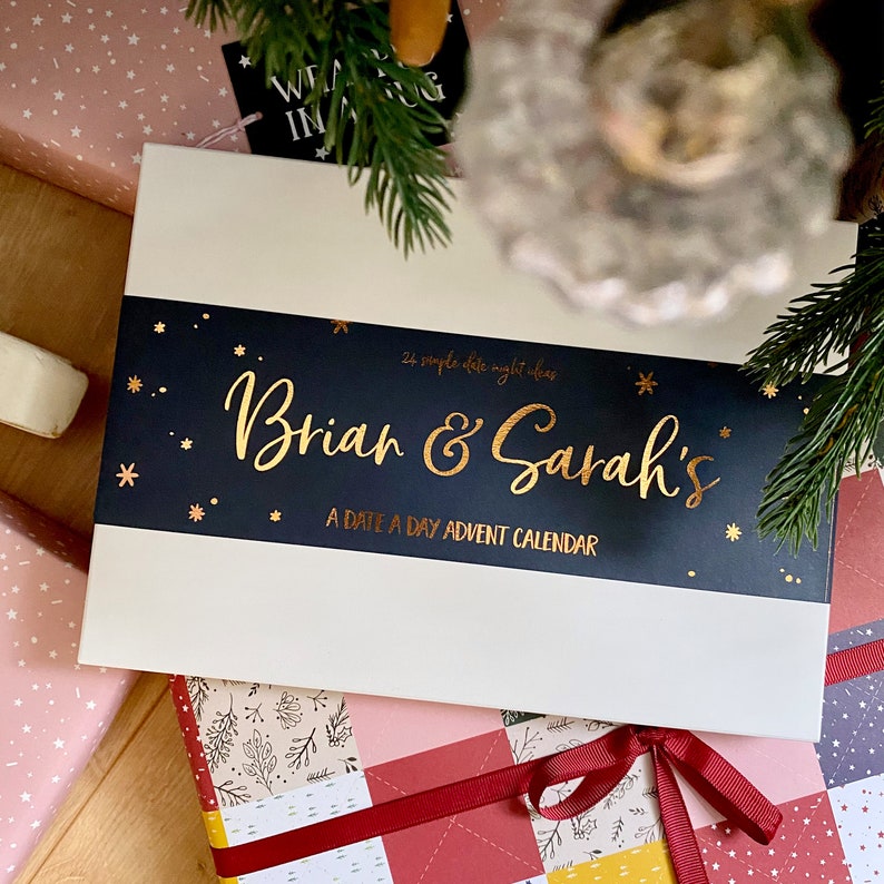 A white advent calendar box with a navy blue sleeve and foiled wording rests on a wooden floor, surrounded by wrapped gifts.