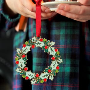 A child wearing a tartan dress holds a vintage-style enamel Christmas wreath ornament with green holly leaves and red berries, hanging from a red ribbon, alongside a small white candleholder with a lit candle.
