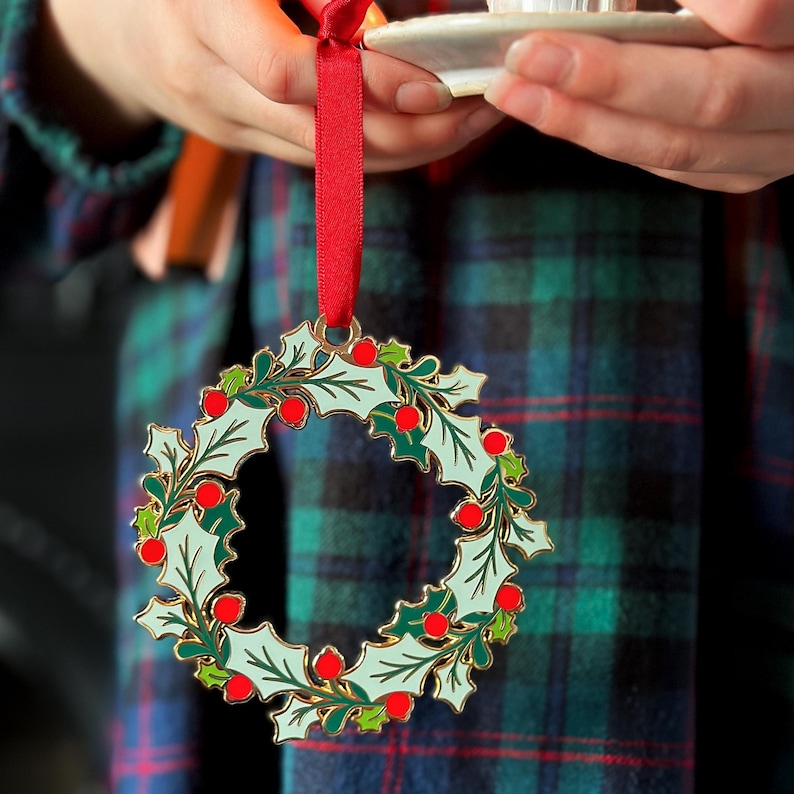 A child wearing a tartan dress holds a vintage-style enamel Christmas wreath ornament with green holly leaves and red berries, hanging from a red ribbon, alongside a small white candleholder with a lit candle.