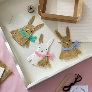 May include: Three felt bunny finger puppets, one white with a pink scarf, one brown with a blue scarf, and one brown with a teal scarf. The puppets are on a white surface with a wooden craft stick and black thread.