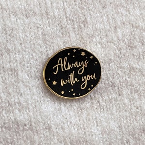 Black oval pin badge with gold ring and small gold stars reading always with you in a gold handwriting font on a cream knitted jumper.