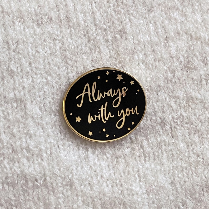 Black oval pin badge with gold ring and small gold stars reading always with you in a gold handwriting font on a cream knitted jumper.