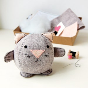 Hand sewn grey felt kitten is placed in front of a craft kit box filled with felt, cotton and stuffing.