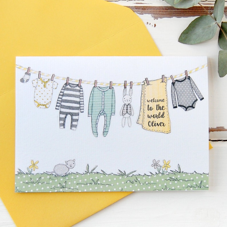 A personalised new baby greetings card with a watercolour painted illustration of a washing line full of baby clothes, lays on a yellow envelope. Both are on a white desk.