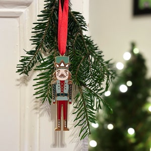 A gold, red, and green enamel Nutcracker Christmas tree decoration hangs from a red ribbon on a door handle, in front a Christmas tree branch.