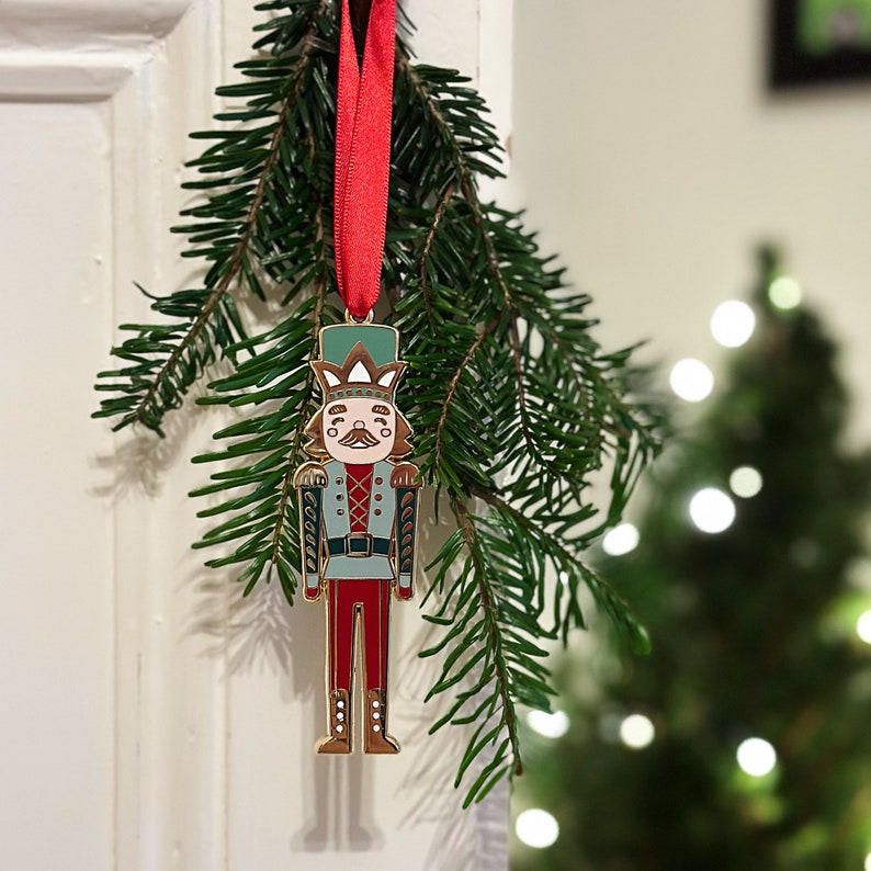 A gold, red, and green enamel Nutcracker Christmas tree decoration hangs from a red ribbon on a door handle, in front a Christmas tree branch.