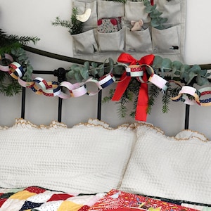 Traditional patchwork design paper chains are strung along the head of a bed along with greenery and red ribbons. An advent calendar hangs above the Christmas paper chains and cosy winter bedding is seen below.