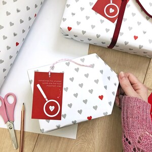 Tiny Heart Valentines Gift Wrap. White gift wrap featuring tiny grey and red hearts, some with tiny messages in is used to wrap a gift and finished off with a red bow.