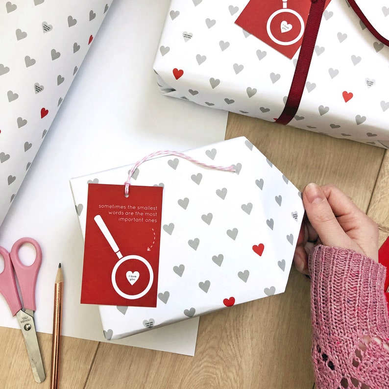 Tiny Heart Valentines Gift Wrap. White gift wrap featuring tiny grey and red hearts, some with tiny messages in is used to wrap a gift and finished off with a red bow.