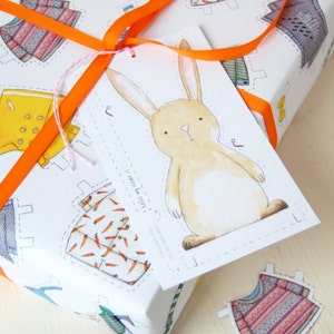 Puede incluir: Un papel de regalo blanco con una ilustraci&oacute;n de un conejo marr&oacute;n y el texto "hello my name is..." en una etiqueta. El papel de regalo presenta un patr&oacute;n repetitivo de recortes de ropa coloridos.
