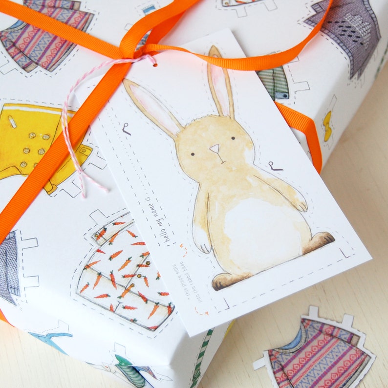 Puede incluir: Un papel de regalo blanco con una ilustraci&oacute;n de un conejo marr&oacute;n y el texto "hello my name is..." en una etiqueta. El papel de regalo presenta un patr&oacute;n repetitivo de recortes de ropa coloridos.