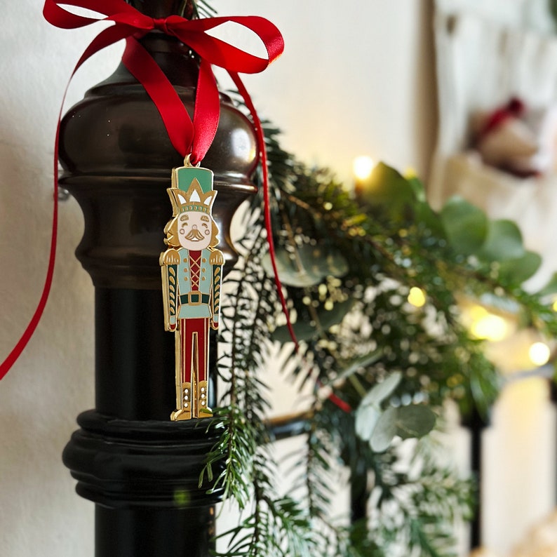 A gold, red, and green enamel Nutcracker Christmas tree decoration hangs from a red ribbon on a door handle, on a pile of wrapped Christmas gifts.