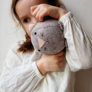 A brunette little girl who is wearing an ivory coloured blouse is holding a hand sewn kitten in her hands, she is tilting the kitten on it's side