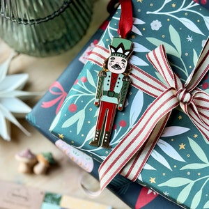 A gold, red, and green enamel Nutcracker Christmas tree decoration hangs from a red ribbon on a door handle, on a pile of wrapped Christmas gifts.