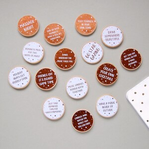 15 wooden disks scattered on a grey background. 7 of the wooden disks have white stickers with copper foiled writing on them, 8 of the disks have copper stickers with white writing on them