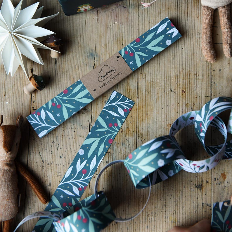 A child is making a set of traditional Christmas paper chains on a wooden table. Christmas decorations lay beside the botanical themed paper chains.