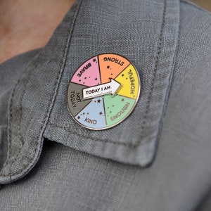 Parent wearing a multicoloured pin badge with an error spinner pointing at different words reading strong, hopeful, enough, kind, not today and brave