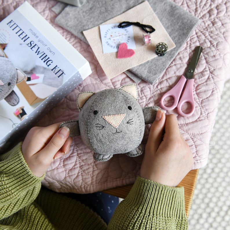 Kitten sewing kit is placed on a pink blanket next to different coloured felt pieces, two metal cotton spools and a pair of pink scissors. There is a person holding a round felt grey kitten in their hands. The person is wearing a green jumper.