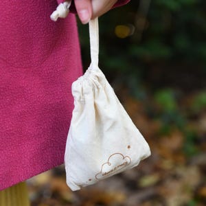 May include: A small, off-white drawstring bag with a cloud and star design, and the text "clara & macy" in a brown script. The bag is held by a person wearing a pink coat. The background is blurred, suggesting an outdoor setting.