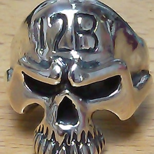 May include: A silver skull ring with the number "123" engraved on the top. The skull has a detailed design with a prominent jawline and a textured surface.