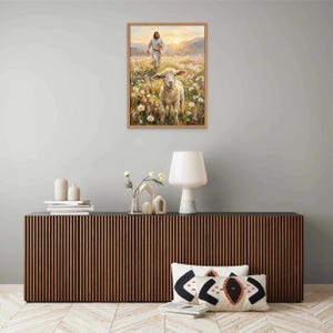 K&ouml;nnte beinhalten: Ein gerahmter Druck zeigt ein Lamm in einem Feld mit Wildblumen, mit einer Figur im Hintergrund. Das Kunstwerk hat eine warme Farbpalette, wobei das Lamm im Mittelpunkt steht. Der Druck wird &uuml;ber einem modernen Holzschrank ausgestellt.
