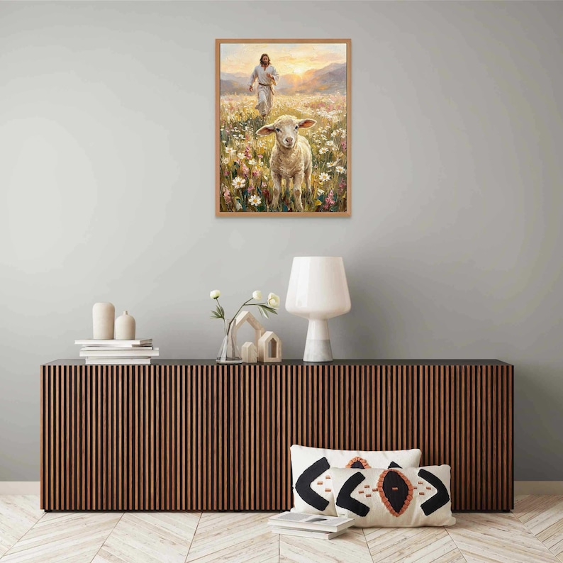 K&ouml;nnte beinhalten: Ein gerahmter Druck zeigt ein Lamm in einem Feld mit Wildblumen, mit einer Figur im Hintergrund. Das Kunstwerk hat eine warme Farbpalette, wobei das Lamm im Mittelpunkt steht. Der Druck wird &uuml;ber einem modernen Holzschrank ausgestellt.
