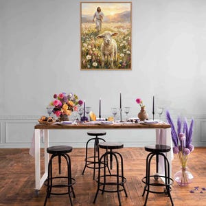 K&ouml;nnte beinhalten: Ein gerahmtes Gem&auml;lde zeigt ein Lamm in einem Blumenfeld mit einer Figur im Hintergrund. Ein langer Holztisch ist mit Kerzen, Blumen und Glaswaren gedeckt. Schwarze Metallhocker mit runden Sitzen sind um den Tisch herum aufgestellt.