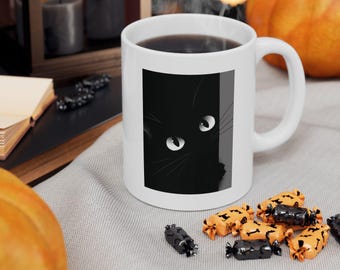 May include: White ceramic mug featuring a black cat graphic with glowing eyes. The mug is filled with a dark liquid, possibly coffee or tea. Halloween-themed candies are scattered around the mug.