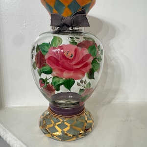 May include: A clear glass vase featuring a hand-painted pink rose with green leaves on the body. The neck has an orange and teal diamond pattern with a dark grey fabric bow. The base is decorated with a teal and gold diamond grid design.