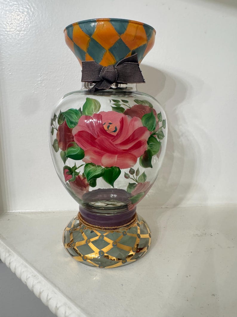May include: A clear glass vase featuring a hand-painted pink rose with green leaves on the body. The neck has an orange and teal diamond pattern with a dark grey fabric bow. The base is decorated with a teal and gold diamond grid design.