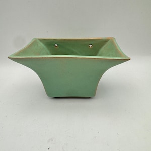 May include: A square-shaped ceramic wall planter with a matte light green glaze. The planter features a wide, flared rim and two small circular holes near the top edge for mounting purposes. The interior and exterior surfaces show a consistent finish.
