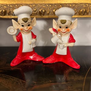 May include: Two ceramic chef figurines. Each figurine is wearing a white chef's hat, a red jacket, and white apron. One holds a small plate, the other a pitcher. The figurines have elf-like ears and rosy cheeks.