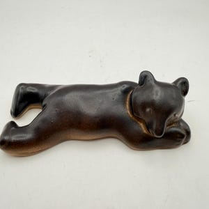 May include: A dark brown ceramic figurine of a bear lying down in a resting position. The bear has a smooth, glossy finish with darker shading along its back and limbs, and lighter brown tones near the base, featuring rounded ears and a snout.